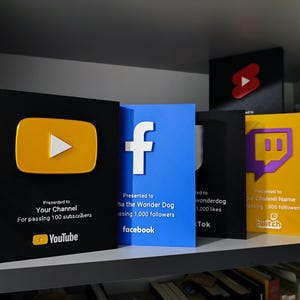 Pode incluir: Uma coleção de prêmios de plataformas de mídia social, incluindo YouTube, Facebook, TikTok e Twitch. Os prêmios são pretos, azuis e amarelos com logotipos e texto indicando marcos de assinantes ou seguidores. Eles são exibidos em uma prateleira.