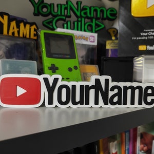 Op de afbeelding: Een wit, rood en zwart 3D-geprint bord met het YouTube-logo en de tekst "YourName".