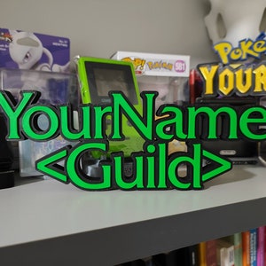 Pode incluir: Um sinal verde e preto impresso em 3D com o texto "YourName <Guild>" em uma prateleira com outros itens.