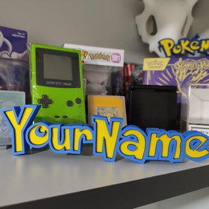 以下が含まれることがあります： 漫画のフォントで「Your Name」と書かれた、青と黄色の3Dプリントされたサイン。サインは、ゲームボーイカラー、ポケモンカード、その他のゲーム関連の記念品が置かれた棚の上に置かれています。