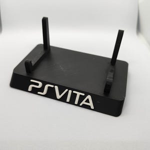 Op de afbeelding: Een zwarte plastic standaard met twee verticale steunen voor een PS Vita handheld-spelconsole. De standaard heeft de tekst "PS VITA" in wit op de voorkant.