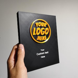 Op de afbeelding: Zwarte plaquette met een gouden cirkel met de tekst "YOUR LOGO HERE" en de tekst "Your Custom text Here" onder de cirkel.