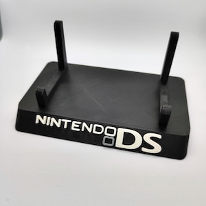 May include: A black plastic stand for a Nintendo DS handheld games console. The stand has two vertical supports and the words "Nintendo DS" printed on the base.