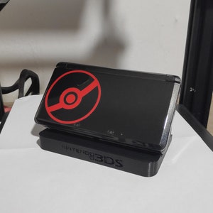 May include: A black Nintendo 3DS handheld gaming console with a red Pokeball logo on the top screen. The console is sitting on a black stand.