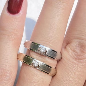 Puede incluir: Dos anillos, uno plateado y otro dorado, cada uno con un pequeño diamante cuadrado. Los anillos están en el dedo de una persona, con el anillo plateado encima. El fondo es una superficie blanca.