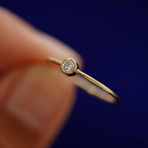 May include: A delicate gold ring featuring a small, round, clear diamond. The ring's band is thin and polished, with the diamond set in a bezel setting. The ring is shown against a dark blue background.