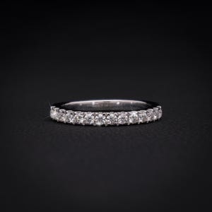 May include: A silver band ring set with numerous small, clear gemstones. The ring is displayed on a black surface, with the gemstones catching the light. The ring is a classic design, suitable for various occasions.