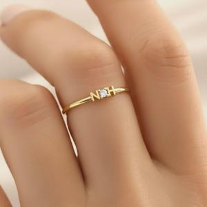 May include: A delicate gold ring featuring the initials "N" and "H" with a small, clear gemstone in the center. The ring is thin and elegant, designed for everyday wear. The ring is shown on a person's finger.