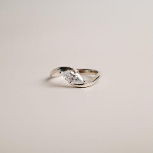 Könnte beinhalten: Ein silberner Ring mit einem Diamanten im Marquise-Schliff. Das Band ist geschwungen und elegant gestaltet. Der Ring ist vor einem neutralen Hintergrund platziert, wodurch der Diamant hervorgehoben wird.