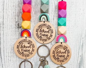 May include: Three wooden keychains with rainbow charms and colorful silicone beads. Each keychain features a round wooden tag with the text "School Nurse" and a list of positive attributes. The keychains have silver key rings with colorful tassels.