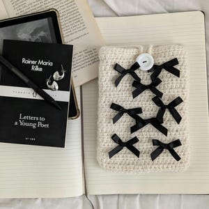 May include: A cream-coloured crocheted tablet sleeve decorated with black satin bows and a white button fastening. A book titled "Letters to a Young Poet" and a pen are visible.