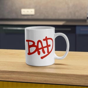 Puede incluir: Taza de cer&aacute;mica blanca con la palabra "BAD" en rojo, con letras estilizadas. La taza tiene un asa curva y est&aacute; colocada sobre una superficie de madera. El fondo muestra una cocina.