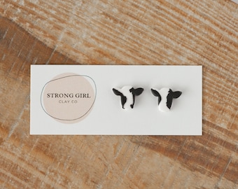 Puede incluir: Un par de pendientes de botón con forma de cabeza de vaca en blanco y negro sobre una tarjeta blanca con el texto "STRONG GIRL CLAY CO" y un logotipo circular. Los pendientes se muestran sobre una superficie de madera.