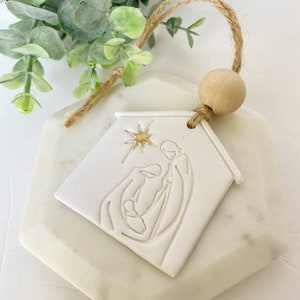 Puede incluir: Adorno de cer&aacute;mica blanca con forma de casa con una estrella dorada y un dibujo lineal sencillo de la Sagrada Familia en su interior. El adorno est&aacute; sujeto a una cuerda de yute con una cuenta de madera.