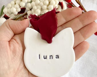 Puede incluir: Un adorno blanco de cerámica con forma de gato con el nombre "luna" en letras negras. El adorno tiene una cinta roja oscura para colgar. Bayas blancas decorativas y bayas rojas en el fondo.