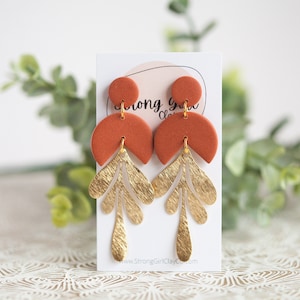 May include: A pair of terracotta and gold dangle earrings. The earrings feature a half-circle shape with a gold leaf design. The earrings are on a white card with the text "Strong Girl Clay".