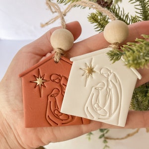Puede incluir: Dos adornos navide&ntilde;os de cer&aacute;mica que representan la escena de la natividad. Un adorno es rojo y el otro es blanco. Ambos adornos presentan una estrella y una representaci&oacute;n simple de Mar&iacute;a, Jos&eacute; y el ni&ntilde;o Jes&uacute;s.