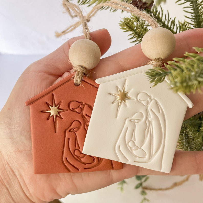 Puede incluir: Dos adornos navide&ntilde;os de cer&aacute;mica que representan la escena de la natividad. Un adorno es rojo y el otro es blanco. Ambos adornos presentan una estrella y una representaci&oacute;n simple de Mar&iacute;a, Jos&eacute; y el ni&ntilde;o Jes&uacute;s.