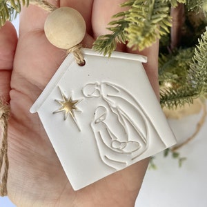 Puede incluir: Adorno de cer&aacute;mica blanca con forma de casa con una estrella dorada y una imagen en relieve de la escena de la natividad en el interior. El adorno est&aacute; colgado de una cuerda de yute marr&oacute;n.