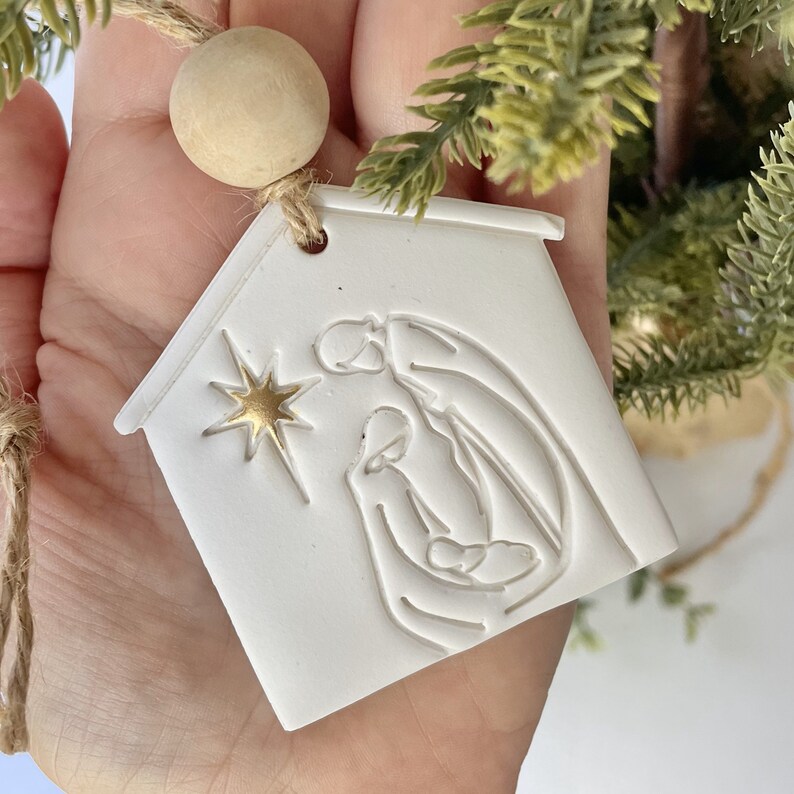 Puede incluir: Adorno de cer&aacute;mica blanca con forma de casa con una estrella dorada y una imagen en relieve de la escena de la natividad en el interior. El adorno est&aacute; colgado de una cuerda de yute marr&oacute;n.