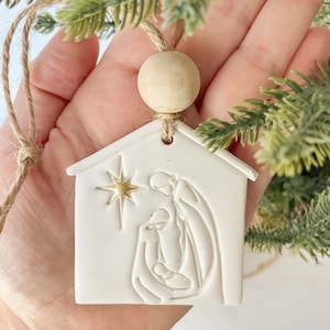Puede incluir: Adorno de cer&aacute;mica blanca con forma de casa con una estrella dorada y una imagen en relieve de la escena de la natividad en el interior. El adorno est&aacute; colgado de una cuerda de yute marr&oacute;n con una cuenta de madera.