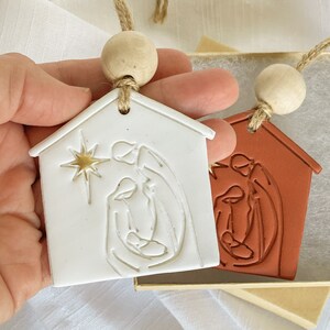 Puede incluir: Dos adornos navide&ntilde;os de cer&aacute;mica con forma de casas con una escena de la natividad en su interior. Un adorno es blanco con detalles dorados y el otro es de terracota.