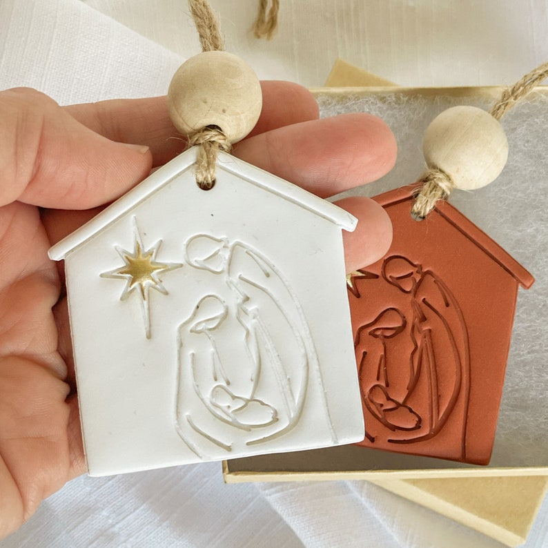 Puede incluir: Dos adornos navide&ntilde;os de cer&aacute;mica con forma de casas con una escena de la natividad en su interior. Un adorno es blanco con detalles dorados y el otro es de terracota.