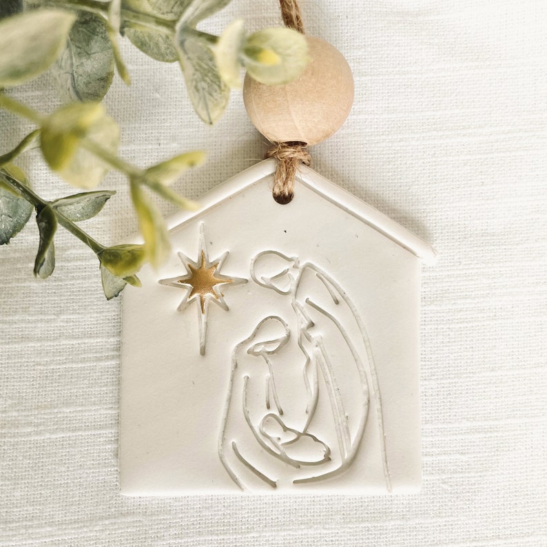Puede incluir: Adorno de cer&aacute;mica blanca con forma de casa con una estrella dorada y una silueta blanca de la escena de la natividad en su interior. El adorno est&aacute; colgado de una cuerda de yute marr&oacute;n con una cuenta de madera.
