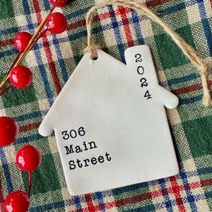 May include: A white ceramic house-shaped ornament with the numbers "2024" and the address "306 Main Street" printed in black. The ornament hangs from a natural twine loop. Red berries and a plaid background complete the scene.