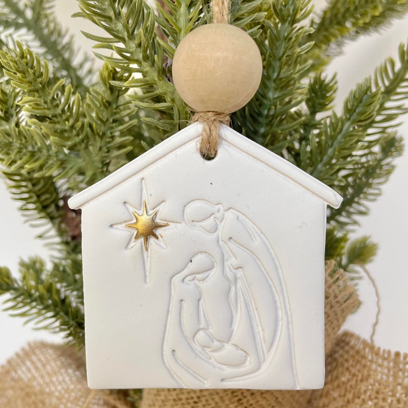 Puede incluir: Adorno navide&ntilde;o de cer&aacute;mica blanca con forma de casa con una estrella dorada y una silueta blanca de la escena de la natividad en el interior. El adorno est&aacute; colgado de una cuerda de yute marr&oacute;n con una cuenta de madera.