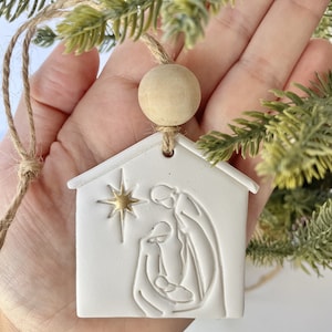 Puede incluir: Adorno de cer&aacute;mica blanca con forma de casa con una estrella dorada y una imagen en relieve de la escena de la natividad en el interior. El adorno est&aacute; colgado de una cuerda de yute marr&oacute;n con una cuenta de madera.