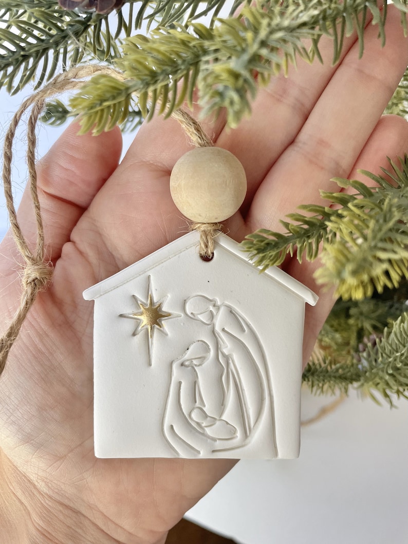 Puede incluir: Adorno de cer&aacute;mica blanca con forma de casa con una estrella dorada y una imagen en relieve de la escena de la natividad en el interior. El adorno est&aacute; colgado de una cuerda de yute marr&oacute;n con una cuenta de madera.