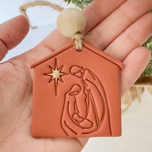 Puede incluir: Un adorno navide&ntilde;o de color terracota con forma de casa con una estrella dorada y una escena de la natividad en su interior. El adorno est&aacute; colgado de una cuerda de yute con una cuenta de madera.