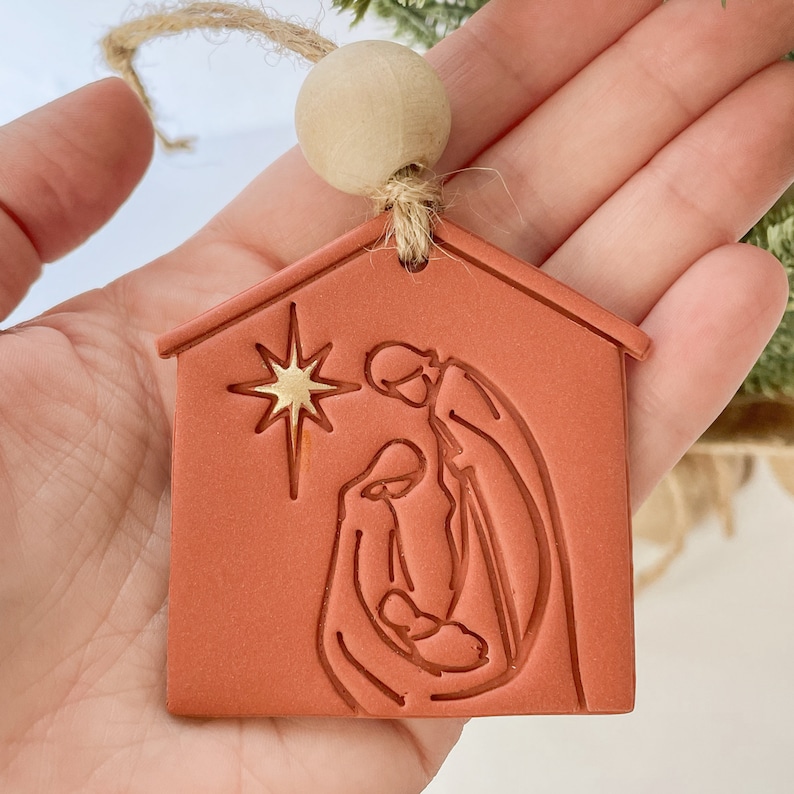 Puede incluir: Un adorno navide&ntilde;o de color terracota con forma de casa con una estrella dorada y una escena de la natividad en su interior. El adorno est&aacute; colgado de una cuerda de yute con una cuenta de madera.