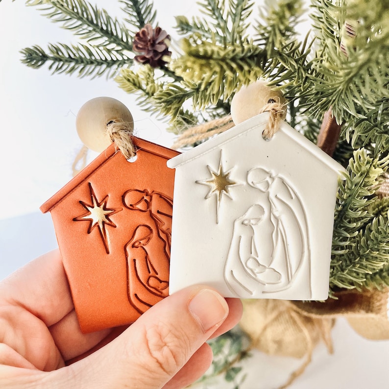 Puede incluir: Dos adornos navide&ntilde;os de cer&aacute;mica con forma de casas con una escena de la natividad en su interior. Los adornos son blancos y naranjas con una estrella dorada en cada uno.