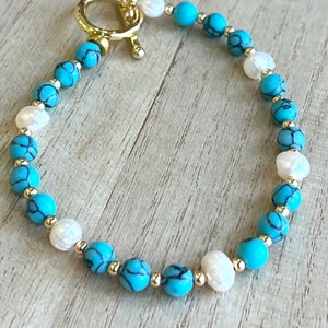 May include: A turquoise and white beaded bracelet with a gold clasp. The bracelet is made with small, round beads and has a simple, elegant design.