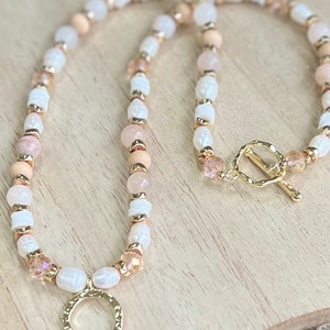 May include: A long, delicate necklace made of white, pink, and brown beads with a gold circle pendant. The necklace has a gold clasp.