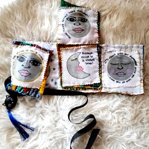 May include: A fabric accordion-style book with four white panels featuring hand-painted moon faces and text. The panels have gold-stitched edges and colorful fabric borders. A black ribbon and a small black cat charm with a blue tassel are attached.