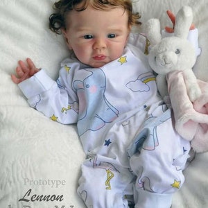 Puede incluir: Una mu&ntilde;eca realista de beb&eacute; con cabello casta&ntilde;o rizado que lleva un mameluco blanco con un estampado de elefante azul y arco&iacute;ris. La mu&ntilde;eca sostiene un juguete de peluche blanco en forma de conejo. El texto "Prototype Lennon by Dawn McLeod" es visible en la imagen.