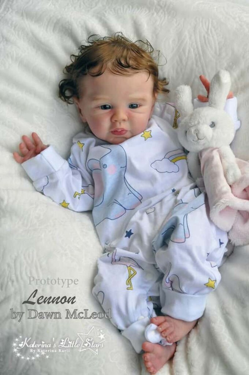 Puede incluir: Una mu&ntilde;eca realista de beb&eacute; con cabello casta&ntilde;o rizado que lleva un mameluco blanco con un estampado de elefante azul y arco&iacute;ris. La mu&ntilde;eca sostiene un juguete de peluche blanco en forma de conejo. El texto "Prototype Lennon by Dawn McLeod" es visible en la imagen.