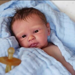 Puede incluir: Un mu&ntilde;eco beb&eacute; realista con cabello casta&ntilde;o y ojos azules, envuelto en una manta azul claro y sosteniendo un chupete.