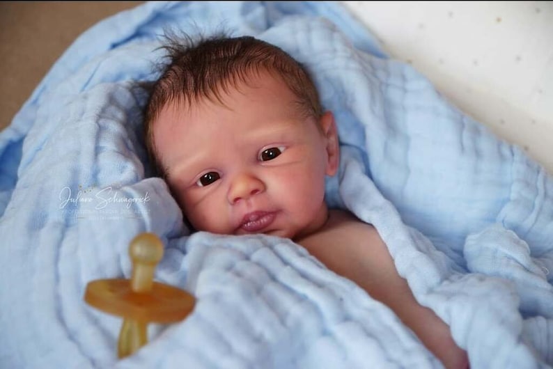 Puede incluir: Un mu&ntilde;eco beb&eacute; realista con cabello casta&ntilde;o y ojos azules, envuelto en una manta azul claro y sosteniendo un chupete.