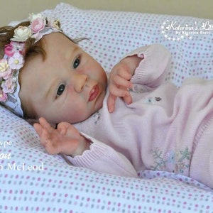 Puede incluir: Una mu&ntilde;eca de beb&eacute; de punto rosa con una diadema de flores, tumbada sobre una manta de lunares blancos y rosas. La mu&ntilde;eca est&aacute; etiquetada como "Prototype Lemour by Dawn McLeod".