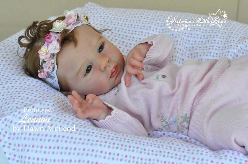 Puede incluir: Una mu&ntilde;eca de beb&eacute; de punto rosa con una diadema de flores, tumbada sobre una manta de lunares blancos y rosas. La mu&ntilde;eca est&aacute; etiquetada como "Prototype Lemour by Dawn McLeod".