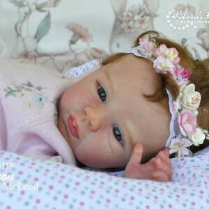 Puede incluir: Primer plano de una mu&ntilde;eca beb&eacute; durmiendo con una corona de flores. La mu&ntilde;eca tiene ojos azules, mejillas sonrosadas y un su&eacute;ter de punto rosa. La mu&ntilde;eca est&aacute; acostada sobre una manta de lunares blancos y rosas. El texto "Prototype Lennon by Dawn McLeod" es visible en la imagen.