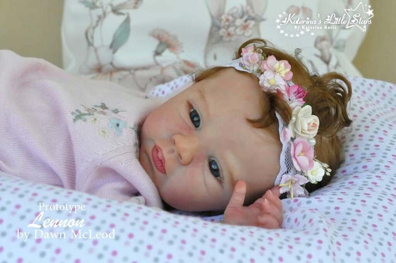 Puede incluir: Primer plano de una mu&ntilde;eca beb&eacute; durmiendo con una corona de flores. La mu&ntilde;eca tiene ojos azules, mejillas sonrosadas y un su&eacute;ter de punto rosa. La mu&ntilde;eca est&aacute; acostada sobre una manta de lunares blancos y rosas. El texto "Prototype Lennon by Dawn McLeod" es visible en la imagen.