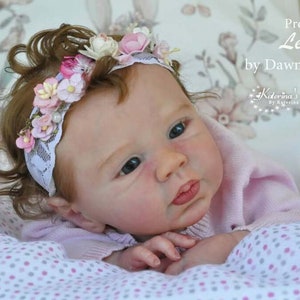 Puede incluir: Primer plano de una mu&ntilde;eca reborn con una diadema de flores rosas. La mu&ntilde;eca tiene ojos azules y lleva un su&eacute;ter rosa. La mu&ntilde;eca est&aacute; tumbada sobre una manta blanca y rosa con lunares.