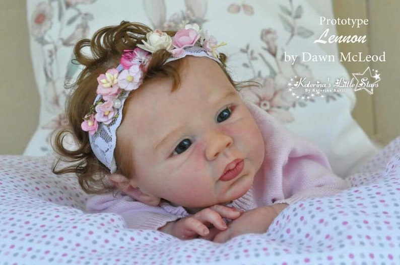 Puede incluir: Primer plano de una mu&ntilde;eca reborn con una diadema de flores rosas. La mu&ntilde;eca tiene ojos azules y lleva un su&eacute;ter rosa. La mu&ntilde;eca est&aacute; tumbada sobre una manta blanca y rosa con lunares.