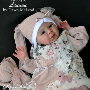 Puede incluir: Una mu&ntilde;eca de beb&eacute; rosa y blanca con un vestido floral rosa y blanco, un gorro rosa con orejas de conejo y un conejito de crochet rosa y blanco. La mu&ntilde;eca est&aacute; tumbada sobre una manta rosa y blanca. El texto "Prototype Lennon by Dawn McLeod" es visible en la esquina superior izquierda de la imagen. El texto "Katerina's Little Stars by Katerina Karli" es visible en la esquina inferior izquierda de la imagen.