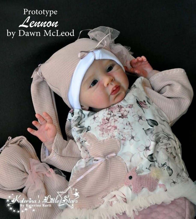 Puede incluir: Una mu&ntilde;eca de beb&eacute; rosa y blanca con un vestido floral rosa y blanco, un gorro rosa con orejas de conejo y un conejito de crochet rosa y blanco. La mu&ntilde;eca est&aacute; tumbada sobre una manta rosa y blanca. El texto "Prototype Lennon by Dawn McLeod" es visible en la esquina superior izquierda de la imagen. El texto "Katerina's Little Stars by Katerina Karli" es visible en la esquina inferior izquierda de la imagen.